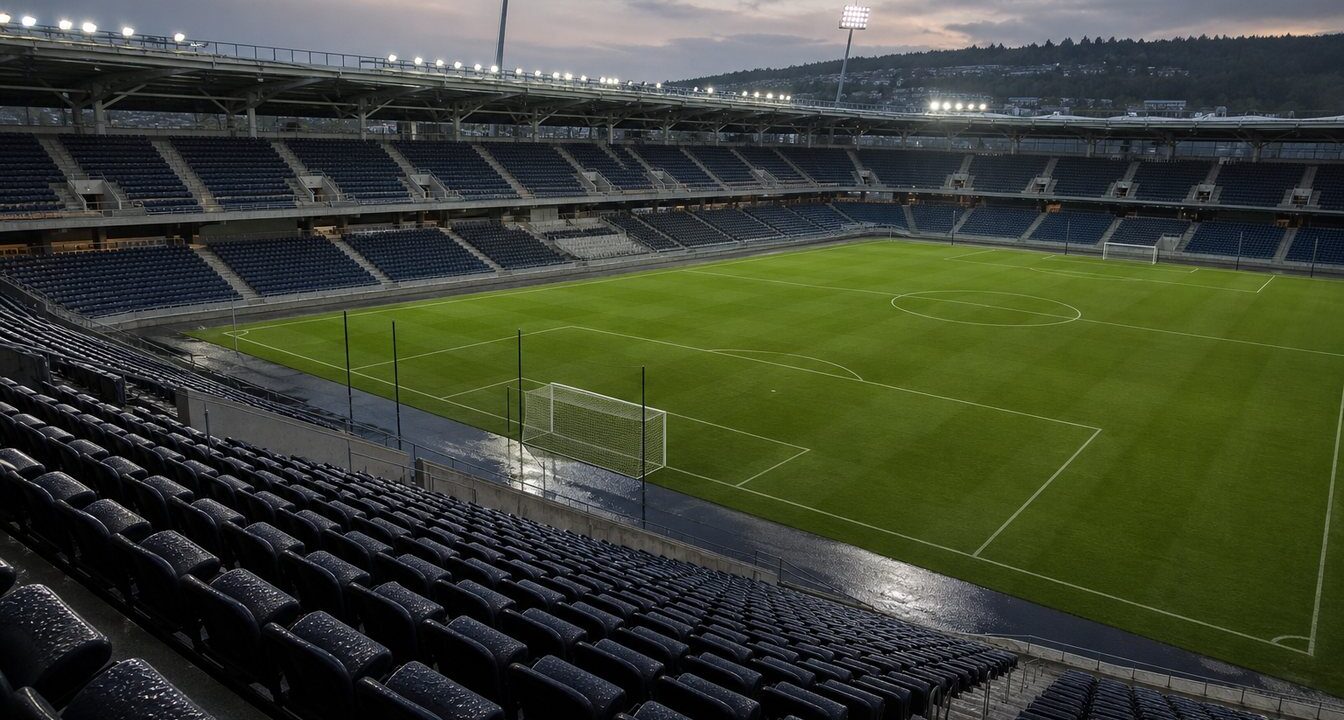 Tom fotbollsarena i skymningen med tända strålkastare och regnvåta läktare